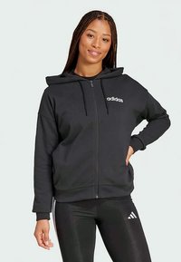 Junge Frau mit langen Zöpfen, die einen schwarzen Adidas-Hoodie mit Reißverschluss und schwarze Leggings mit Adidas-Logo trägt, lächelt und hat eine Hand auf der Hüfte.
