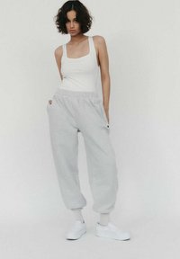 Młoda kobieta stoi na jednolitym tle, ubrana w białą koszulkę na ramiączkach, jasnoszare dresy, mając ręce w kieszeniach, w białych skarpetkach i białych trampkach.