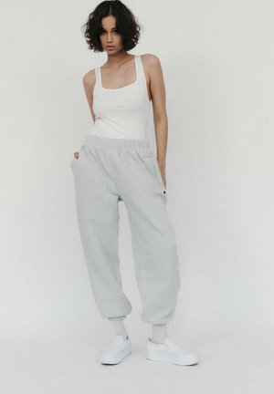 Młoda kobieta stoi na jednolitym tle, ubrana w białą koszulkę na ramiączkach, jasnoszare dresy, mając ręce w kieszeniach, w białych skarpetkach i białych trampkach.