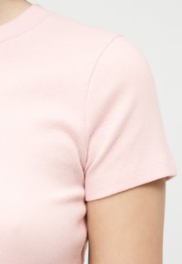 Κοντινό πλάνο ενός ανοιχτό ροζ πουκαμίσου με κοντά μανίκια πάνω στο άνω μέρος του κορμού και του χεριού ενός ατόμου, πάνω σε μια απλή ανοιχτή φωτεινή επιφάνεια.