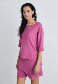 Ροζ σετ loungewear που περιλαμβάνει κοντομάνικη, φαρδιά μπλούζα με ραβδωτή υφή και αντίστοιχες σορτς. Ο σχεδιασμός περιλαμβάνει μια πλευρική σχισμή και διακριτική λεπτομέρεια branding.