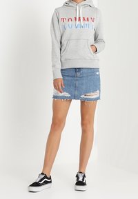 Kobieta modelka w szarej bluzie z kapturem z napisem "TOMMY", w podartej dżinsowej spódnicy mini i czarno-białych trampkach, stojąca na białym tle.