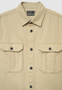 Beiges Hemd mit regulärer Passform, das einen spitzen Kragen, schwarze Knöpfe und zwei Vorderseiten mit Klappenverschlüssen aufweist. Strukturierter Stoff.