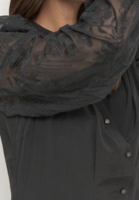 Abito nero con maniche trasparenti ricamate con motivi floreali, chiusura frontale con bottoni e un corpo liscio e texturizzato.
