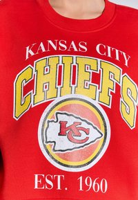 Piros kapucnis pulóver, melyen "KANSAS CITY CHIEFS" felirat található sárga és fehér színben, valamint egy kör alakú logó, amelyen a piros "KC" egy sárga körvonalban látható.
