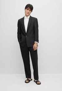 Traje negro con cierre de un solo botón, pantalones a medida a juego y una camisa blanca con cuello. Lleva sandalias de deslizar negras.