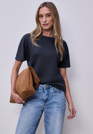 Kvinde med langt blondt hår iført en navyblå kortærmet top og lyseblå jeans, der holder en stor brun foldet clutch-taske.