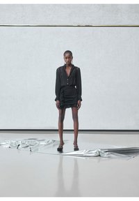 Eine Frau steht auf glänzendem silbernem Stoff, trägt eine schwarze durchsichtige Bluse mit langen Ärmeln und einen gerafften Minirock, vor einem schlichten strukturierten Wandhintergrund.