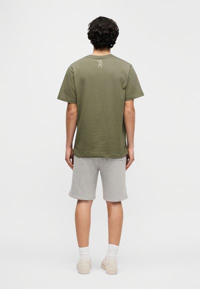 Μακρυμάνικη μπλούζα από βαμβάκι σε ελιά πράσινο χρώμα με κοντά μανίκια και μικρό γραφικό στο πίσω μέρος· συνδυασμένη με γκρι σορτς και ανοιχτόχρωμα αθλητικά παπούτσια.