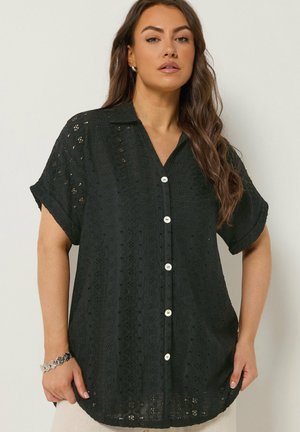 Kvinde iført sort kortærmet skjorte med knapper og hulmønster i blonder samt opkørte ærmer, parret med et sølvarmbånd på venstre håndled.