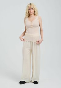 Beige Trägershirt mit Spitzenbesatz und drei Knöpfen, kombiniert mit lockeren, hellen Hosen. Modell trägt schwarze Schuhe vor einem schlichten Hintergrund.