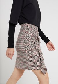 Jupe crayon à carreaux beige, rouge et noir avec un détail de volants sur le côté. Fabriquée en tissu léger, elle présente une silhouette ajustée.