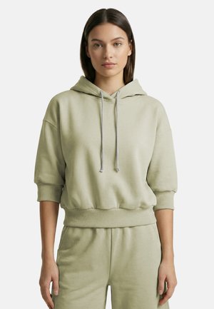 Jonge vrouw draagt lichtgroene cropped hoodie met trekkoorden en bijpassende broek, staand tegen een effen achtergrond.