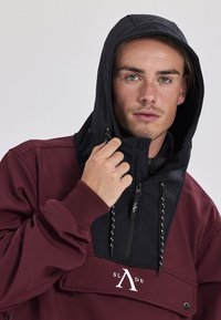 Mørkerød og sort hoodie med høj krave, lynlås foran og justerbar hætte. Har logo og dekorative snore. Glat stofstruktur.