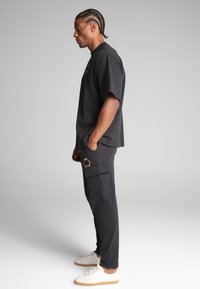 Sort oversized t-shirt med korte ærmer, parret med mørke cargo-bukser med en sidelomme og et guld-emblem. Hvide sneakers fuldender looket.