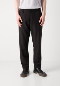 Pantalons noirs fuselés avec taille élastique, fermeture à cordon de serrage, poches latérales et texture lisse. Portés avec des chaussures de sport noires.