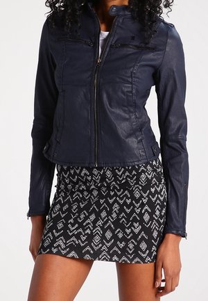 Vrouw die een marineblauwe leren jas met rits draagt en een zwart-witte minirok met een geometrisch patroon, staand met één hand ontspannen langs haar zijde.