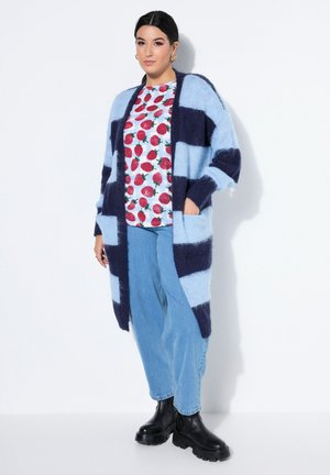 Femme portant un long cardigan rayé bleu et marine sur un t-shirt blanc avec des fraises rouges, un jean bleu clair et des bottes noires épaisses.