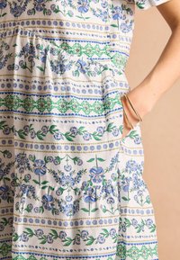 Kvetinové bavlnené šaty s modrými a zelenými vzormi na bielom pozadí, s volánovým lemom a bočnými vreckami.