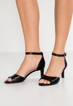 Stopy ubrane w czarne skórzane sandały na średnim obcasie z odkrytymi palcami i paskami na kostkach, stojące na jasnej podłodze.