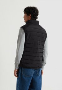 Schwarze Steppweste mit hohem Kragen, schmalen horizontalen Einsätzen und glatter Stoffoberfläche, getragen über einem grauen Langarmshirt und blauen Jeans.