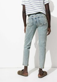 Jeans bleu clair avec un design tapered droit, style cinq poches et délavage. Portés avec une chemise rayée et des chaussures marron à enfiler.