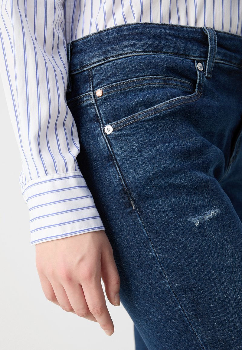 Jeans de mezclilla azul oscuro con una rodilla desgastada, herrajes plateados y una silueta ajustada, combinados con una camisa blanca con rayas azules.