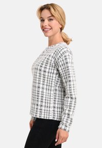 Grau-weißer Karosweater mit langen Ärmeln, Rundhalsausschnitt und strukturierter Oberfläche. Über schwarzen Hosen getragen.