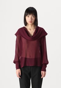 Blusa trasparente bordeaux con un ampio scollo drappeggiato e maniche lunghe a sbuffo, abbinata a pantaloni neri. Tessuto morbido, design fluido.