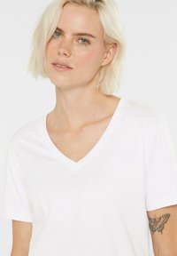 Weißes V-Ausschnitt-T-Shirt aus weichem Stoff, mit kurzen Ärmeln und lässiger Passform. Hautfarbener Hintergrund mit sichtbarem Schmetterlingstattoo.