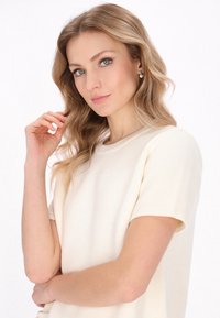 Top a maglia color crema con maniche corte e colletto tondo. Il tessuto presenta una leggera texture e la modella sta toccando delicatamente i capelli.