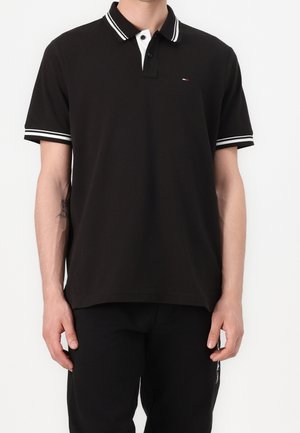 Homme portant un polo noir avec un col blanc et des rayures blanches sur les manches, assorti à un pantalon noir sur un fond blanc uni.