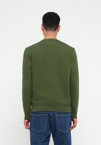 Muž v zelenom dlhom rukáve s jednoduchým dizajnom a modrými džínsami, pohľad zozadu, na bielom pozadí.