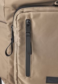 Sac à dos en tissu doux de couleur beige, doté d'une grande poche zippée, de tirettes de fermeture éclair noires, d'une sangle texturée et d'un petit patch logo. Fini lisse.