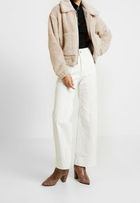 Beige fuskpälskort jacka med krage, svart topp under, högmidjade vita vida byxor och bruna boots med ormskinnsmönster.