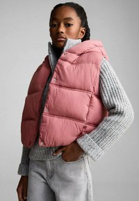 Vest puf roz cu glugă, cu butoane tip clapă, purtat peste un pulover gri, cu model cu nervuri. Modelul are mâinile pe șolduri, evidențiind ținuta.