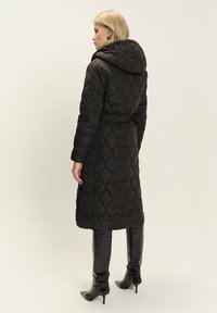 Dlhý čierny prešívaný kabát s kapucňou, s opaskom na páse, textúrovaným diamantovým vzorom a hladkým povrchom. Nosený s čiernymi zaoblenými čižmami po kolená.