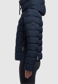 Donkerblauwe gewatteerde jas met capuchon, voorzien van een doorgestikt ontwerp, ritszakken en een logopatch op de bovenarm.