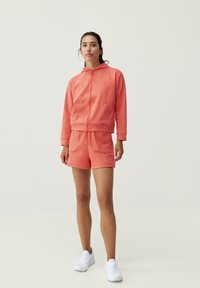 Korallfärgad träningsoutfit bestående av en jacka med dragkedja och fickor samt matchande shorts. Mjukt material med minimalistisk design och vita accenter.