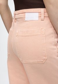 Πίσω όψη ατόμου που φοράει ροζ ανοιχτό χρώμα τζιν με ορατή πίσω τσέπη και λευκή ετικέτα στη μέση, συνδυασμένο με σκούρο καφέ τοπ.