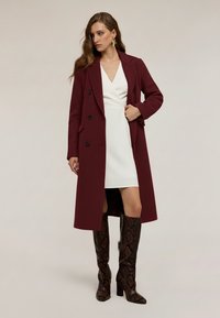 Cappotto bordeaux doppiopetto con revers a lancia, abbinato a un vestito bianco e stivali al ginocchio marroni con stampa pitonata, in piedi su uno sfondo neutro.