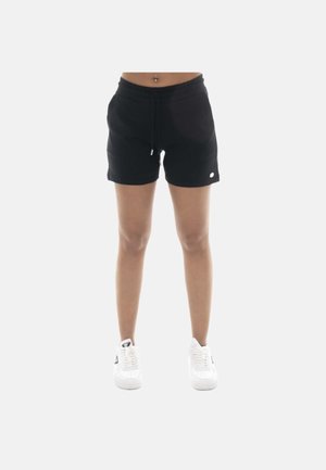 Femme portant un short noir à cordon et des baskets blanches, debout devant un fond clair uni.