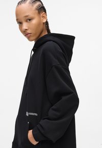 Black hoodie with a large, relaxed fit and a front pocket. Features drawstrings on the hood and embroidered branding on the chest.