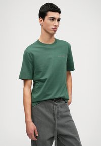 Zaļš kokvilnas t-krekls ar īsām piedurknēm un apaļu kakla izgriezumu, ar mazu gaiši pelēku drukātu logotipu kreisajā pusē.