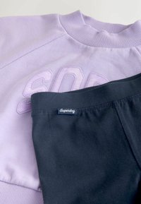 Fialová mikina s vystúpeným nápisom "SOD" a rebrovaným výstrihom, spárovaná s čiernymi legínami označenými "Superdry" na pásku.
