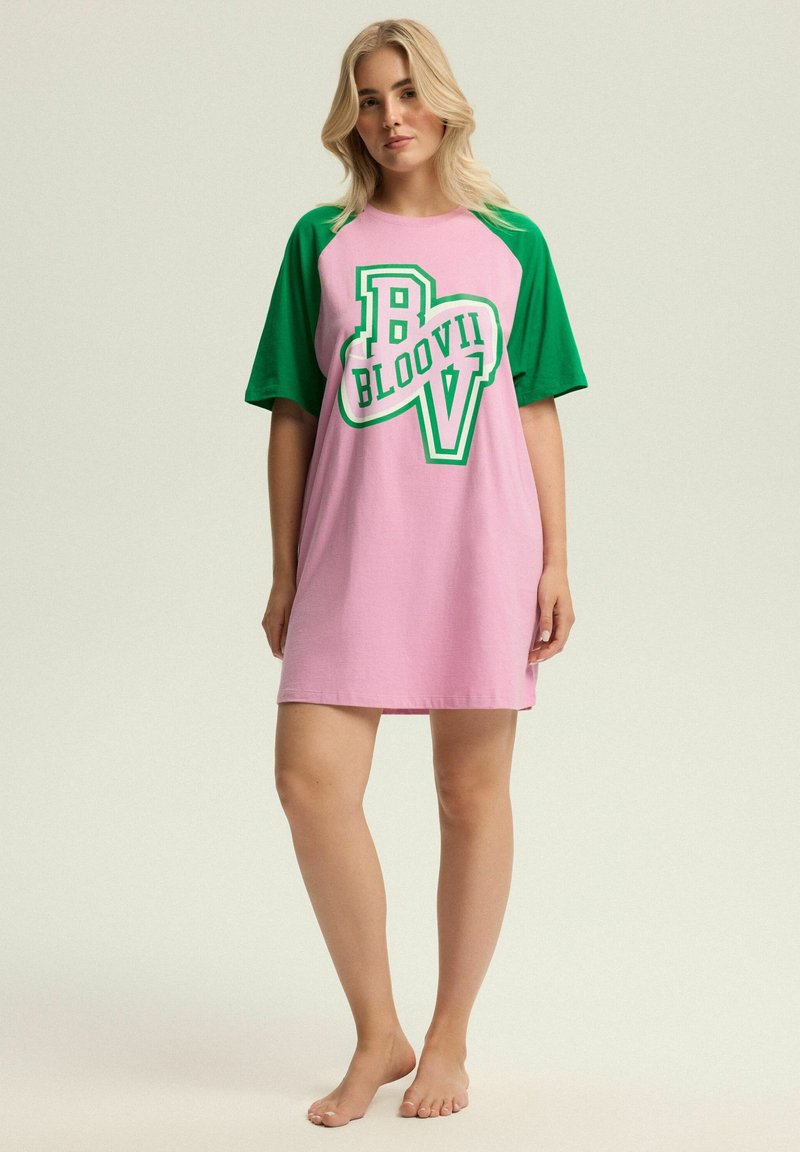 Kobieta stojąca boso, ubrana w różową sukienkę z zielonymi rękawami i zielonym logo "B BLOOVII V" z przodu.