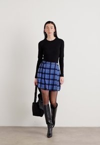 Black long-sleeve top, blue plaid flared skirt, thigh-high black boots, carrying a black tote bag. Smooth fabric with a fitted design.