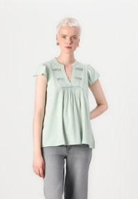 Blusa verde chiaro con scollatura a V, maniche corte ricamate e corpino arricciato con rifiniture in pizzo decorativo. Tessuto morbido.