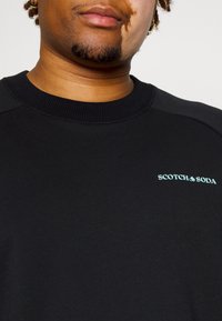 Black sweatshirt with ribbed neckline, featuring a turquoise "SCOTCH & SODA" logo. Made of soft, textured fabric with raglan sleeves.