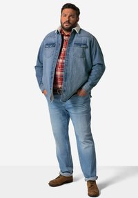 Giacca di denim blu con colletto in shearling, camicia a quadri rossa sotto, jeans azzurri chiari e stivali di pelle marrone. Presenta due tasche sul petto.